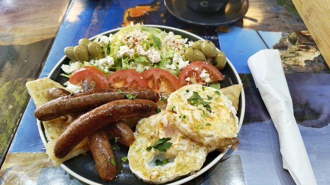 Grilled sausages, fried eggs, tomato, feta, olives and salad on a plate.
