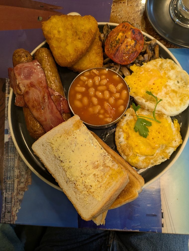 A full English breakfast on a dark plate with eggs, bacon, sausage, hash browns, tomato, beans and toast.