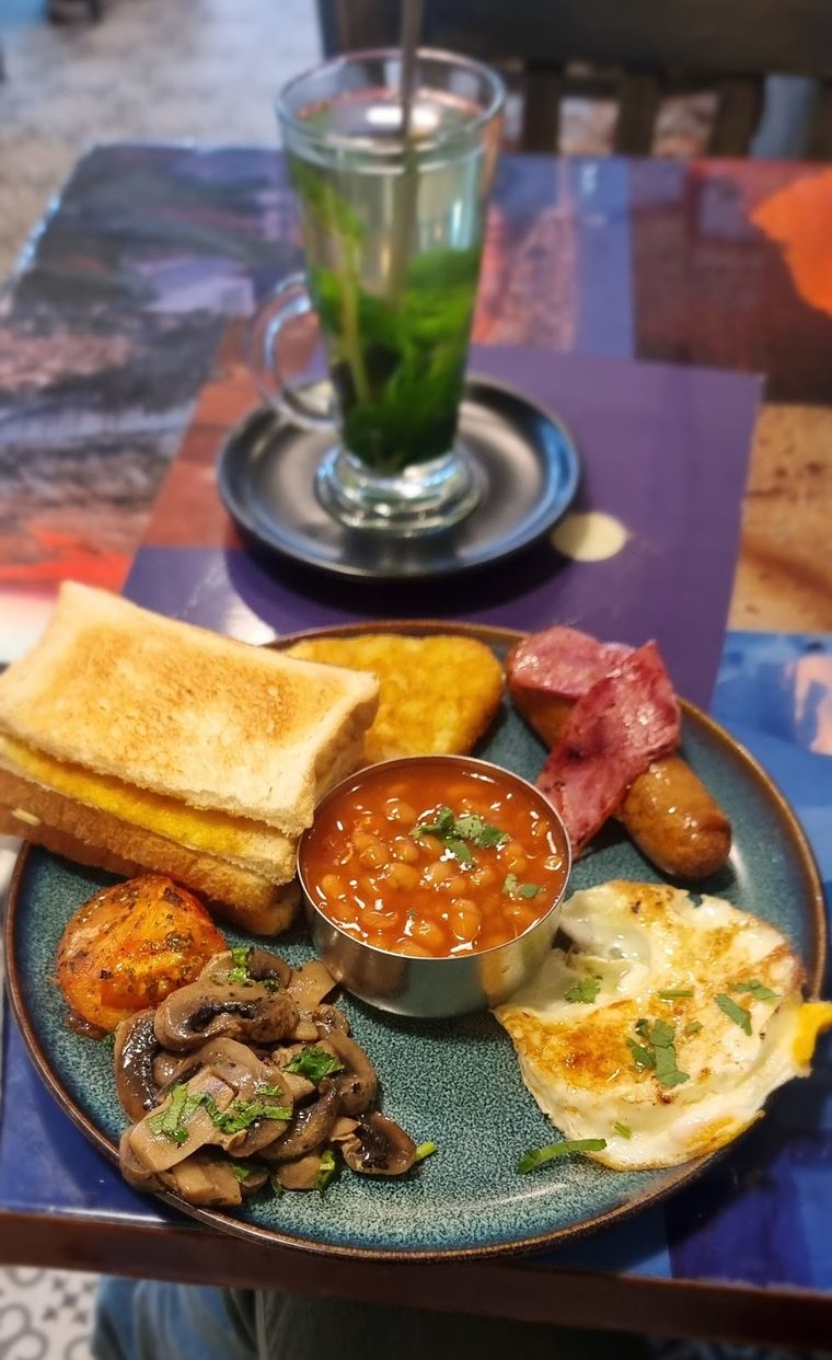 A breakfast plate of fried eggs, mushrooms, sausages, hash browns, beans and toast with a green juice.
